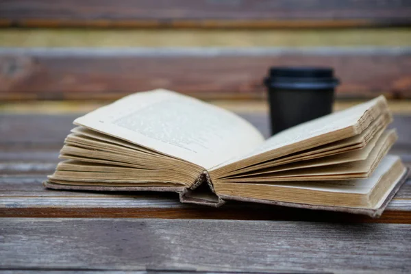 An old shabby thick book lies on a wooden bench next to a cup of — Stock Photo, Image
