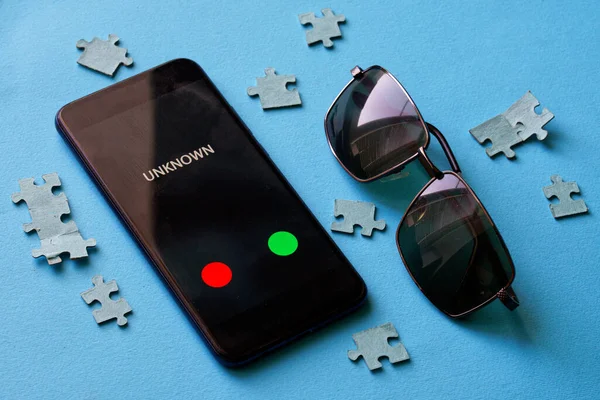 A smartphone with an unknown call lies on a blue surface next to a puzzle and sunglasses. The problem of anonymity and espionage. Protection of personal information