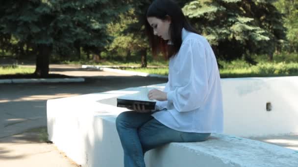 Девушка сидит на фонтане в парке, держит книгу в руках и мечтает . — стоковое видео