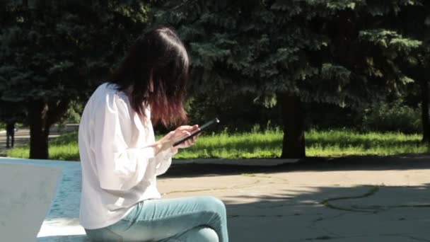 Дівчина сидить на фонтані в парку, грає в гру — стокове відео
