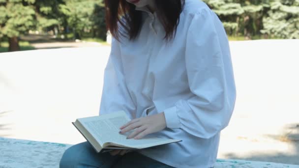 Dziewczyna siedzi i czyta książkę w parku. — Wideo stockowe
