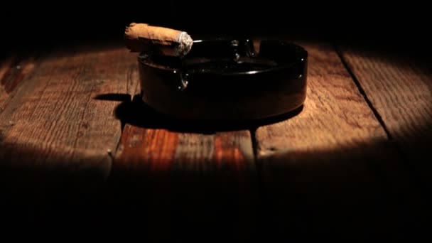 El cigarro está acostado en un cenicero sobre una mesa de madera. Iluminado por el centro de atención . — Vídeo de stock