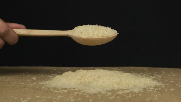 Hand raises a wooden spoon with rice grains. Grain falls from a spoon onto a pile of grains on a black background. — Stock Video