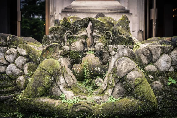 Traditional balinese stone sculpture of a man and woman with moss in garden near pond in Bali, Indonesia.