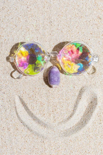 Funny smiling face made of kaleidoscope multicolor glasses and purple sea shell on sand background with copy space, flat lay, vertical composition — Stock Photo, Image