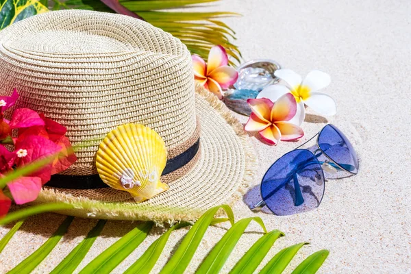 Cappello di paglia beige con occhiali da sole blu, conchiglie colorate, fiori di frangipani e foglie di palma verde sulla sabbia, concetto di vacanza estiva , — Foto Stock