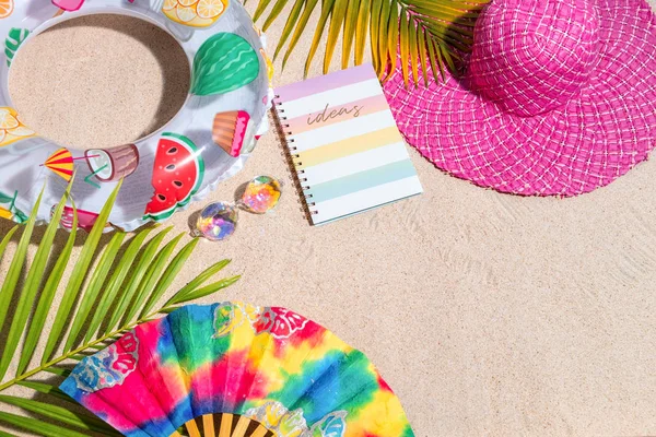 Almohadilla de notas de escritura colorida con bolígrafo rosa y verde sobre arena, rodeada de hojas de palma verde, conchas marinas, sombrero rosa, juguetes inflables. Fondo de playa de verano, plano laico Imagen De Stock