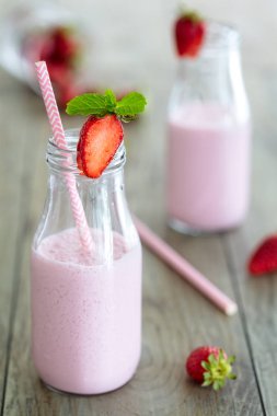 Sağlıklı çilek smoothie, süt shake, bir ahşap arka plan üzerinde dağınık çilek ve pembe saman ile geleneksel şişelemeler içinde. Dikey kompozisyon