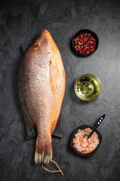 Fresh raw red snapper fish with red pepper, olive oil and Himalayan rock salt on dark background. Top view. Vertical composition.