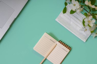 Masaüstünde kalem, dizüstü bilgisayar, maske ve çiçek açan bir elma ağacı dalı olan bir defter. Karantina döneminde evde çalış. Nane rengi arkaplanı