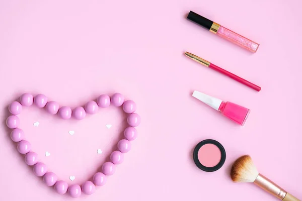 Pembe renkli kızlar için aksesuarlar ve kozmetikler - dudak parlatıcı, dudak kalemi, allık, fırça, tırnak cilası, pembe arka planda pembe kalp şeklinde pembe boncuklar.