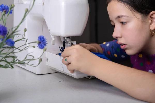 Kız dikiş makinesine mavi şifondan bir elbise dikiyor, dikiş makinesinin yanında vazoda bir çiçek var. Dikiş makinesini kullanan kadın
