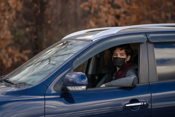 Young man in a face protective mask sits in the blue car on the autumn background. COVID-19 pandemic coronavirus young man wearing surgical mask