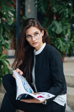 Genç güzel esmer kız, ofis giyim tarzı, bir kafede bir masada sokakta oturan, sokak modeli poz tarzı, bir not defterinotlar yapar, telefonda konuşurken