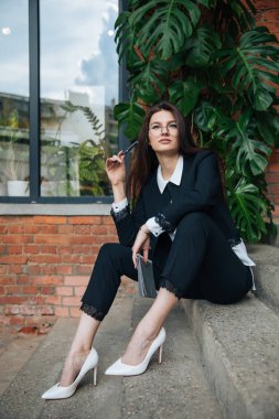 Genç güzel esmer kız, ofis giyim tarzı, bir kafede bir masada sokakta oturan, sokak modeli poz tarzı, bir not defterinotlar yapar, telefonda konuşurken