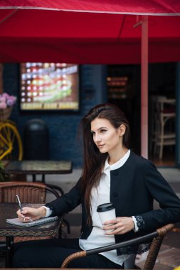 Genç güzel esmer kız, ofis giyim tarzı, bir kafede bir masada sokakta oturan, sokak modeli poz tarzı, bir not defterinotlar yapar, telefonda konuşurken