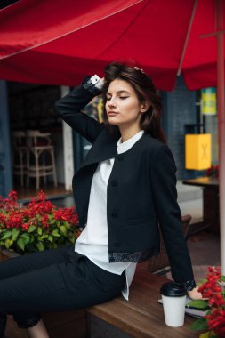 Genç güzel esmer kız, ofis giyim tarzı, bir kafede bir masada sokakta oturan, sokak modeli poz tarzı, bir not defterinotlar yapar, telefonda konuşurken