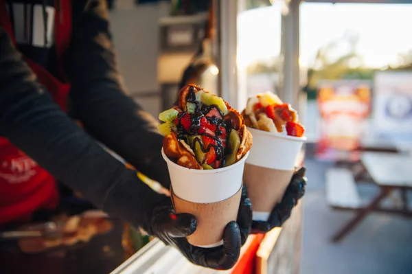 stock image delicious Viennese waffles with fruits chocolate and syrup in a girl's hands in a paper cup, a treat for children and adults