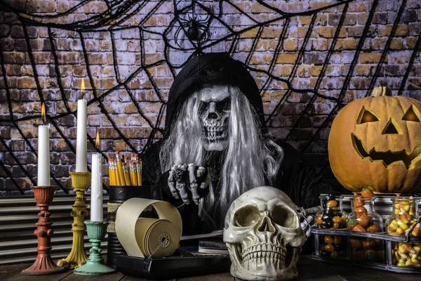 man in Grimm reaper robe scary mask and skeleton hands pointing at camera surrounded by skull jack o lantern burning candles Halloween candy with brick wall background covered with large spider and web