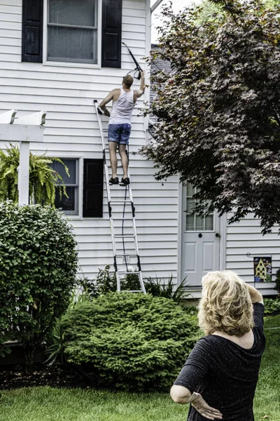 man in shorts on ladder power washing white two story home while woman watches near tree in landscaped back yard on sunny summer day