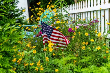 Beyaz çitli ve arka planda mavi Adirondack sandalyesi olan yabani çiçeklerin bahçesinde yatay Amerikan bayrağı