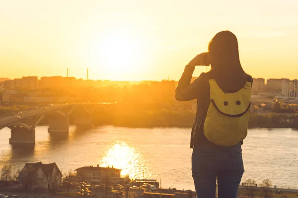 Girl tourist with a backpack stands and takes pictures of the sunset in a golden hour in Nizhny Novgorod city. Picturesque view on city and Kanavinsky bridge across the Oka river. Back view on person