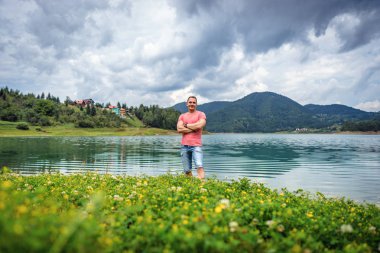 Sırbistan'daki bulutlu gökyüzü altında Zaovine Gölü yaz aylarında ayakta bir genç gülümseyen adam