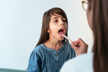 Hastanede küçük kızı muayene eden kadın doktor boğaz ağrısını kontrol ediyor. Çocuk doktoru hastane odasında küçük bir hastanın bademciklerini kontrol ediyor..