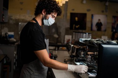 Korona virüsünün yayılmasını önlemek için cerrahi maske ve eldiven takarak kahve yapan genç adam, barista..