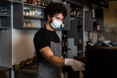 Korona virüsünün yayılmasını önlemek için cerrahi maske ve eldiven takarak kahve yapan genç adam, barista..