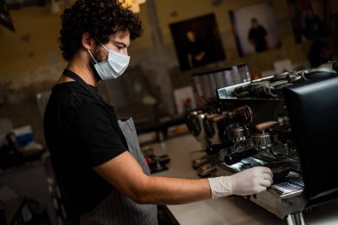 Korona virüsünün yayılmasını önlemek için cerrahi maske ve eldiven takarak kahve yapan genç adam, barista..