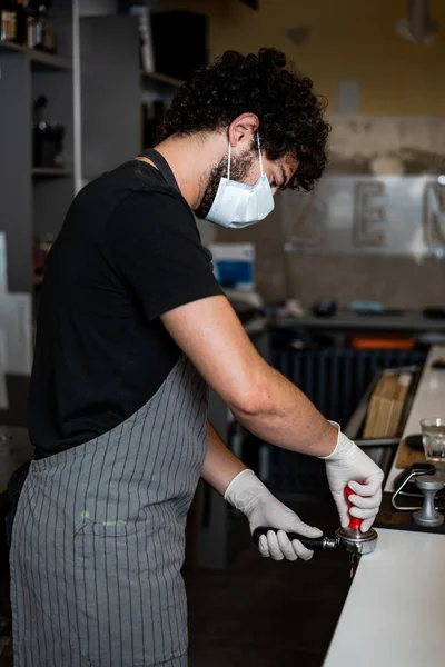 Young man, barista, making coffee espresso while wearing surgical mask and gloves for preventing corona virus spread - Bar safety working concept.