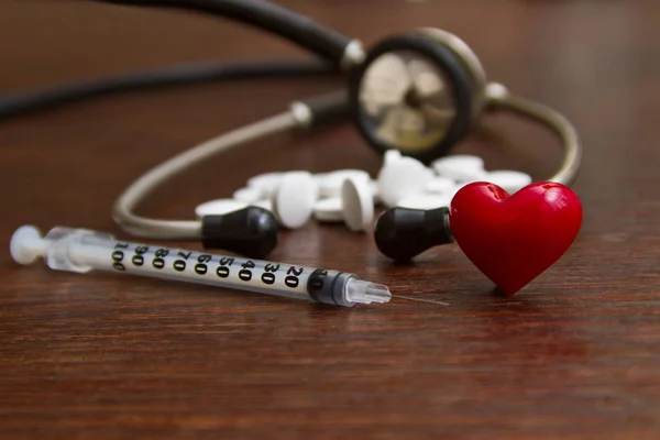 Medical stethoscope and a red heart with a syringe and pills on a wooden table