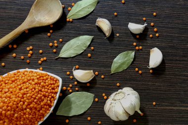 Sarımsak ve üstten görünüm bakan defne yaprakları yanında karanlık bir ahşap zemin üzerine beyaz bir kase kırmızı mercimek.