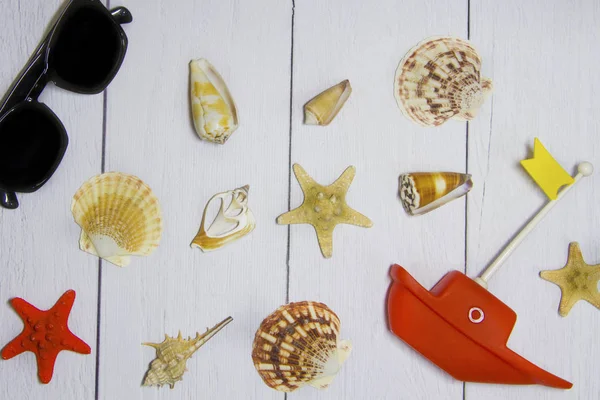 Концепция путешествия - игрушечная лодка, шляпа, солнцезащитные очки, различные экзотические раковины и другие морские сокровища на светлом деревянном фоне с видом на песок сверху — стоковое фото