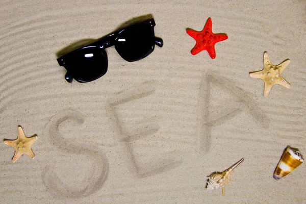 「海」という言葉は、サングラスと貝殻を持つビーチの砂の上に手で書かれています — ストック写真