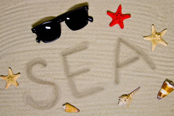 「海」という言葉は、サングラスと貝殻を持つビーチの砂の上に手で書かれています — ストック写真