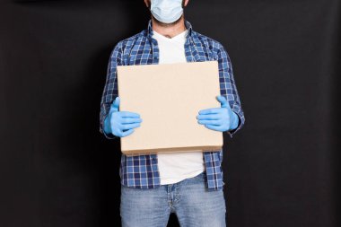 Tıbbi maske takan bir kuryenin elinde karton bir kutu taşıyan kurye ve eldivenli bir servis elemanı koli kutusunu tutuyor..