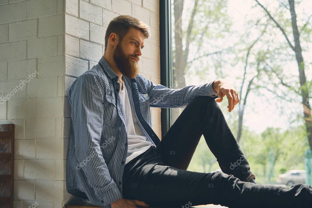 Young attractive bearded man rests and looks out the window. Enjoy the morning afternoon. Healthy lifestyle concept.
