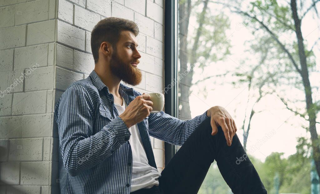 Young attractive bearded man rests and looks out the window. Enjoy the morning afternoon. Healthy lifestyle concept. Drink fresh bracing coffee looking into the distance