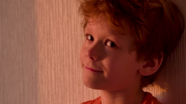 红发卷发的小男孩站在一堵墙在日落的明亮光线的背景上 — 图库视频影像