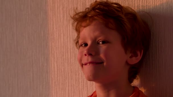 红发卷发的小男孩站在一堵墙在日落的明亮光线的背景上 — 图库视频影像