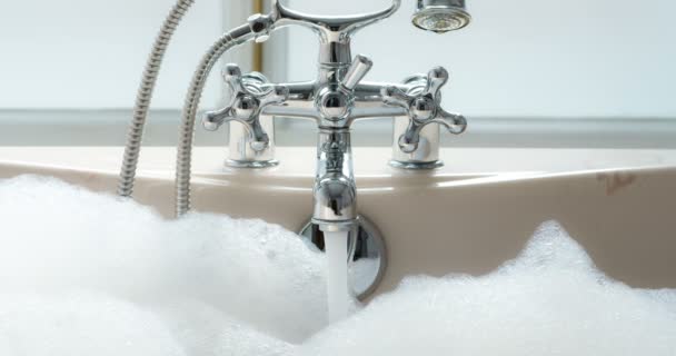 水龙头里的一股水流充满了浴缸 水面上覆盖着丰富的泡沫 女性双手扭动阀门 阻止水流 — 图库视频影像