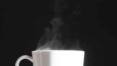 Porselen bardakta sıcak içecek. Işıl ışıl beyaz bir kupa. Siyah arka planda sıcak buhar yavaşça yükseliyor.