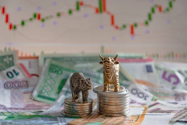 Bronze figures of a bull and a bear near metal coins against the background of paper money and charts. Blur background and perspective. Concept and symbol of stock exchange and stock trading.