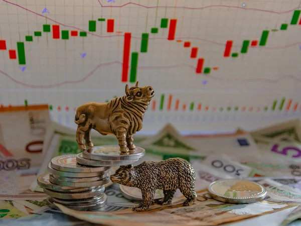 Bronze figures of a bull and a bear near metal coins against the background of paper money and charts. Blur background and perspective. Concept and symbol of stock exchange and stock trading.