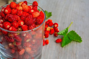 Ahşap bir yüzey üzerinde yaban çilek bir bardak. Yakındaki birkaç çilek ve yaprakları dağınık. Bulanıklık ile arka planda Yaz doğa. Olgun meyveler sağlıklı bir diyet için vitamin açısından zengindir.