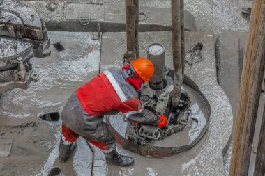 Kırmızı üniformalı, kasklı ve gözlüklü iş delici. O bir asansör yardımıyla bir petrol kuyusu onları kaldırmak ve sondaj devam etmek için matkap boruları asıyor. Çalışan bir kişi kavramı.