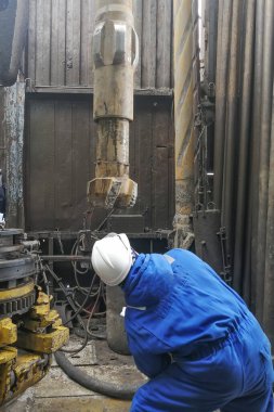Bir matkap ucu uzmanı bir petrol ve gaz kuyusundan çıkarılan parçanın durumunu inceliyor. Mühendis mavi bir tulum ve beyaz bir şapka giyiyor..