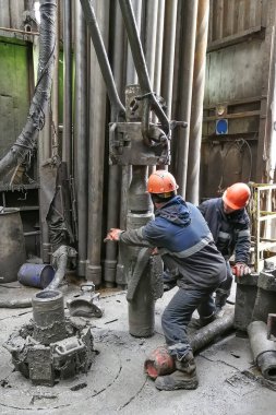 İki sondaj işçisi sondaj ipinin altını söküyor. Spiral kalibratörü söküyorum. Bir makine anahtarıyla çalışmak. Sondaj kulesinin döner platformunda bulunuyorlar..